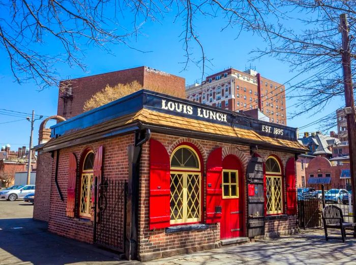 A quaint, red-brick building featuring a bright red door and shutters, with the name Louis’ Lunch displayed above its small shingled roof.