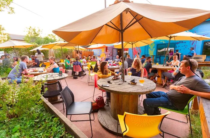 Groups of people enjoying drinks at outdoor patio tables near a brewery.