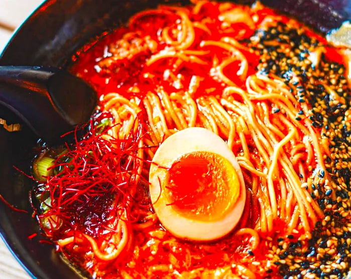 A close-up view of a bowl of ramen featuring squiggly noodles swimming in a rich red broth, topped with an egg, sesame seeds, and various garnishes.