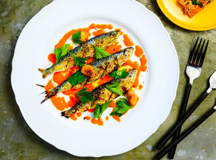 A dish featuring whole grilled sardines topped with orange sauce, parsley, and garlic.