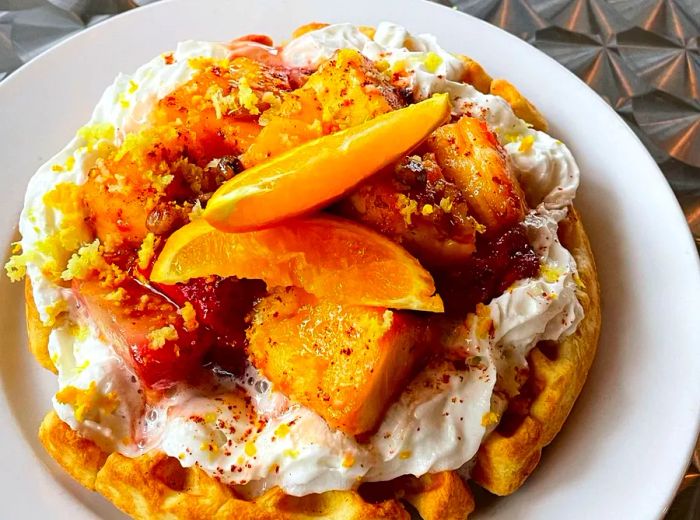 A waffle adorned with cooked fruit, whipped cream, orange slices, and zest.