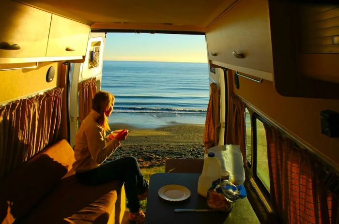 A woman enjoys a cup of coffee while sitting in the open doorway of a campervan, gazing out at a beautiful sea view.