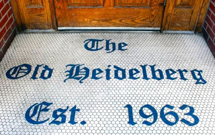 “The Old Heidelberg Est. 1963” is beautifully displayed in tiles on the floor just outside a wooden door.