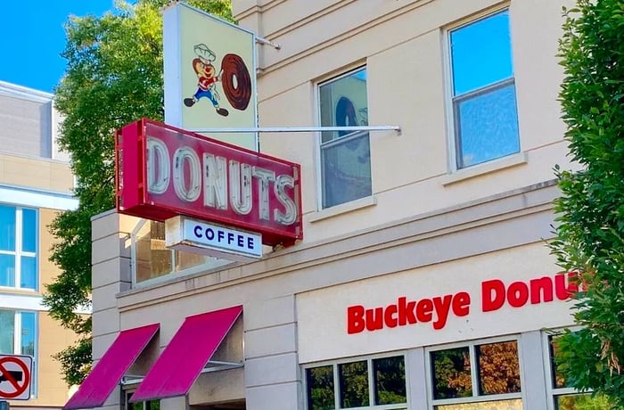 The exterior of the shop features a prominent hanging sign for donuts, another for coffee, and bold lettering showcasing the shop's name. Additionally, there's a whimsical illustration of a chef-like animal character pushing a giant donut.