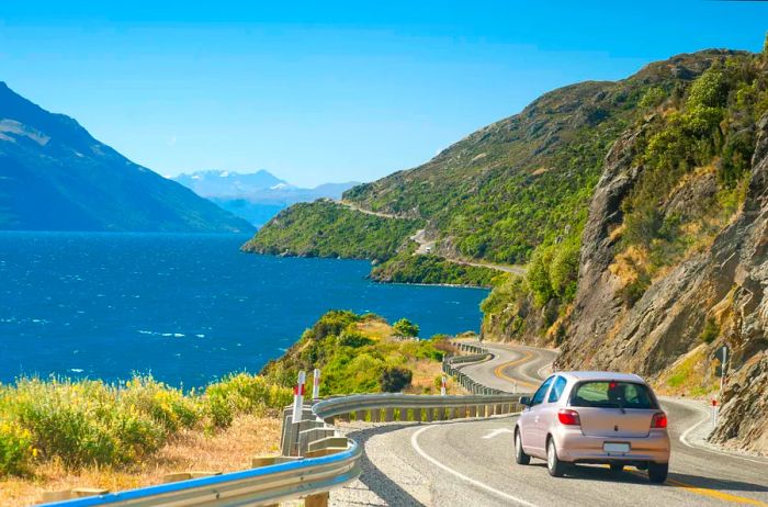 A small gray car drives along a two-lane highway in New Zealand, with expansive hills visible in the background.