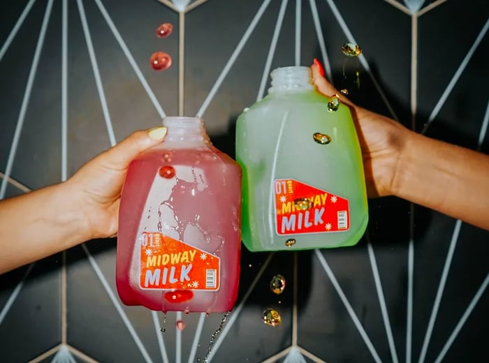 Two hands toast with small milk jugs labeled Midway Milk, filled with vibrant red and green beverages.