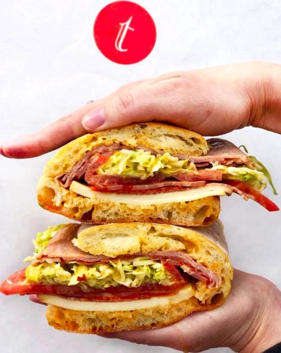 A pair of hands holds a sandwich, beautifully layered with thinly sliced meats and fresh vegetables.