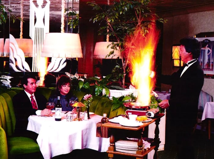 An old photograph capturing diners in a round green booth as they observe a waiter flambéing a dish at their table.