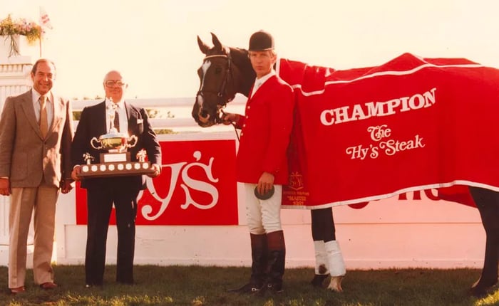 A jockey atop a horse adorned with a red blanket bearing the words Champion The Hy’s Steak stands alongside two men in suits, including Hy Aisenstat, who proudly holds a large trophy.