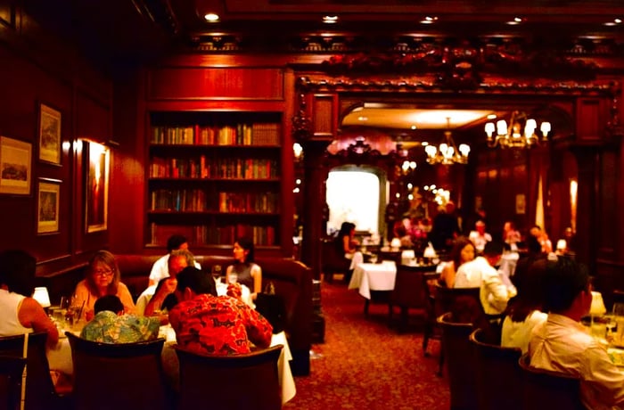 Diners enjoy their meals in elegantly curved wooden chairs at tables draped with white tablecloths, surrounded by bookshelves, chandeliers, and intricately carved woodwork.