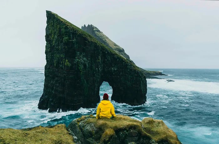 A woman in a yellow raincoat gazes at the Drangarnir arch in the Faroe Islands.