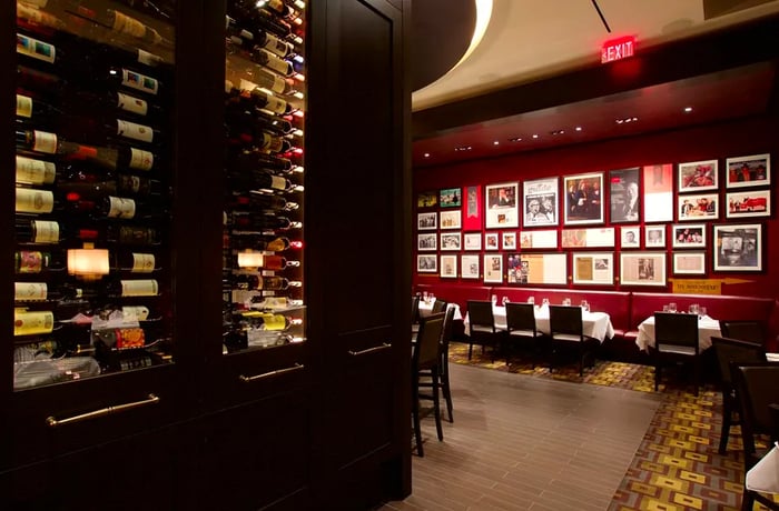 A spacious wine vault features glass displays showcasing bottles, curving towards a seating area adorned with a wall of memorabilia and news clippings.