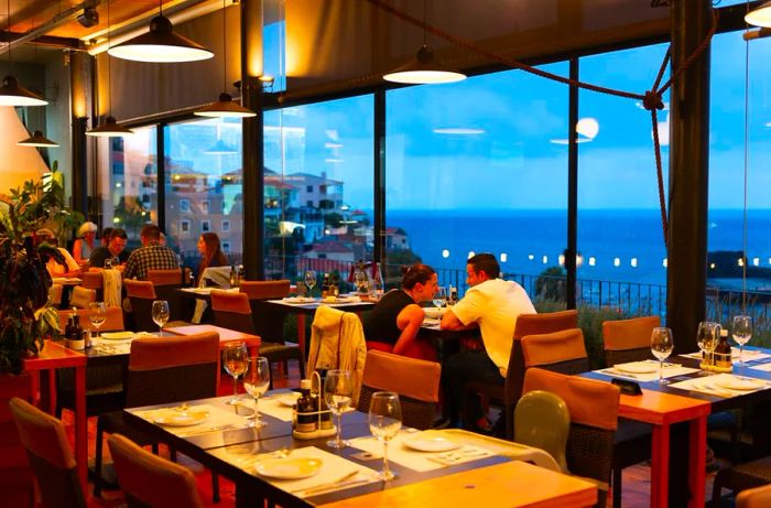 An inviting restaurant interior with a sea view, featuring diners enjoying their meals beneath stylish pendant lighting.