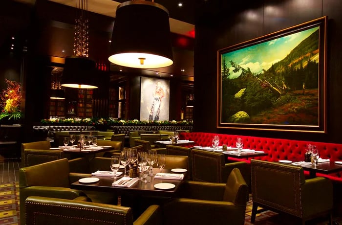 The opulent interior of the steakhouse features plush red banquettes, wide leather chairs, contemporary pendant lighting, and large, vibrant artwork against dark walls.