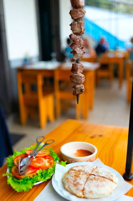 A skewer of meat dangles above a plate of flatbread, accompanied by vegetables, sauce, and tongs.