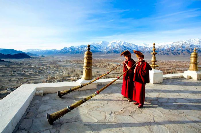 Monks in red robes play horns atop a Ladakh monastery, with majestic mountains in the background.