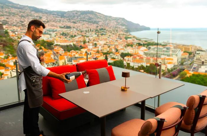 A server fills a glass with Champagne at an empty table on a balcony overlooking a coastal city below.