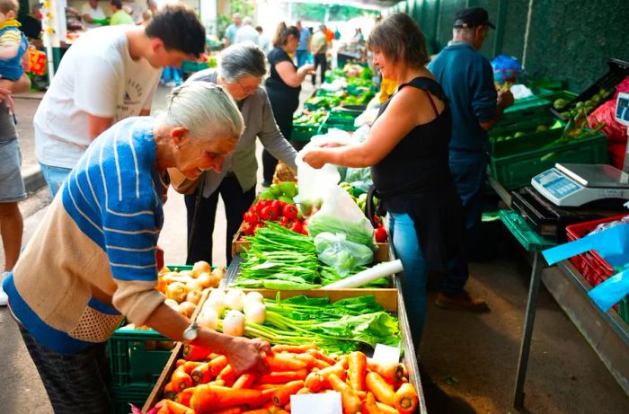 An elderly shopper sifts through a box of carrots at a farmers market.