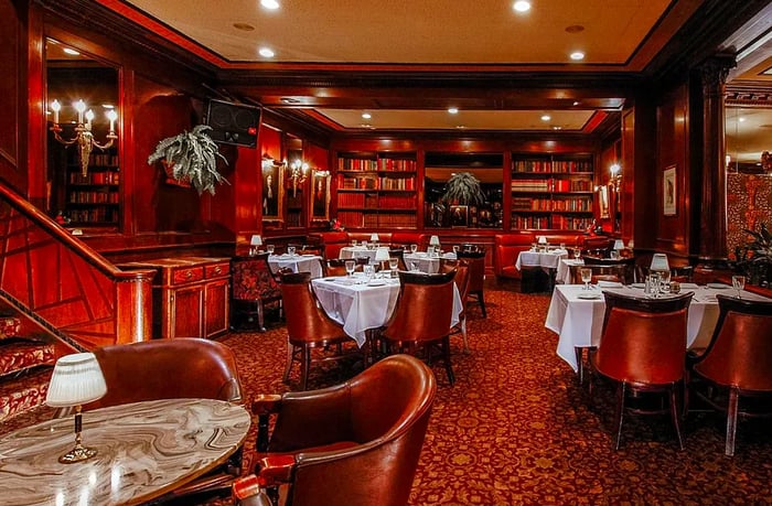 An interior reminiscent of a library, with patterned carpet, curved leather chairs, white tablecloths, ornate sconces, and hanging potted plants.