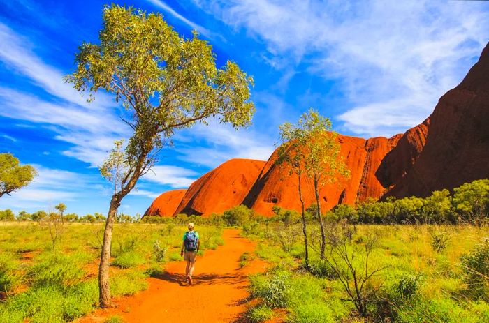 A traveler strolls along a dirt path leading toward the stunning red rocks of Uluru-Kata Tjuta National Park in Australia.