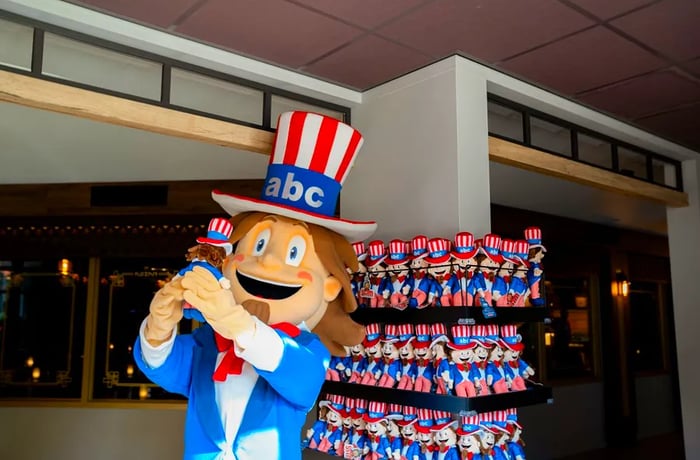 An individual dressed as Uncle Sam holds a small toy replica of himself in front of a display rack filled with more toys.