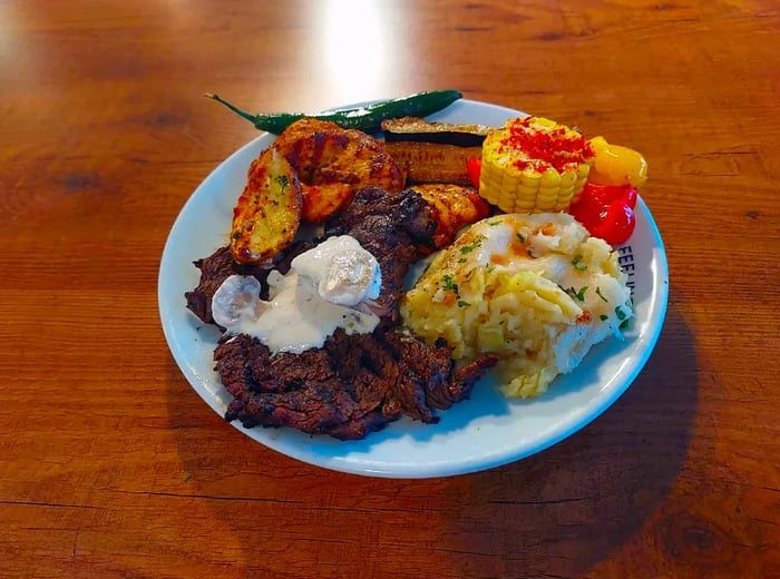 A steak dish adorned with a dollop of creamy butter, accompanied by roasted potatoes, zucchini, corn, bell pepper, and creamy mashed potatoes.