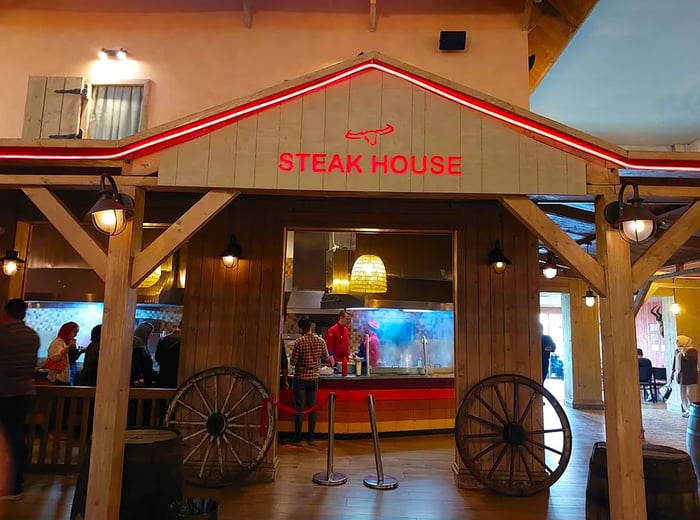 An American West-themed eatery with 'Steak House' emblazoned in red on the exterior, decorated with wagon wheels, and featuring visible chefs working in the kitchen inside.