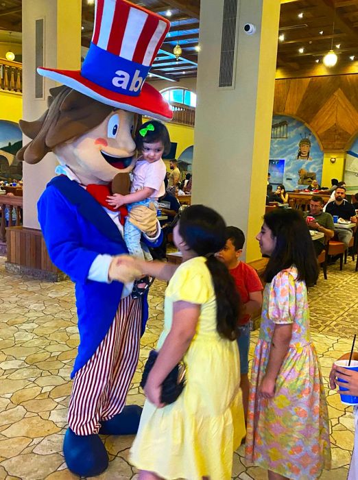 A giant plush Uncle Sam is seen holding a small child while greeting a line of other children in a curved restaurant area.