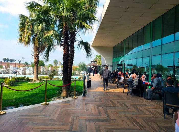 Diners sit at tables next to a tall glass wall, overlooking a lush garden adorned with towering palm trees.