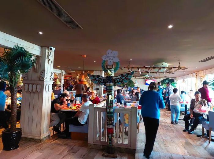 A totem pole labeled Hawaii stands at the entrance of a bustling dining area, adorned with a surfboard and palm trees.