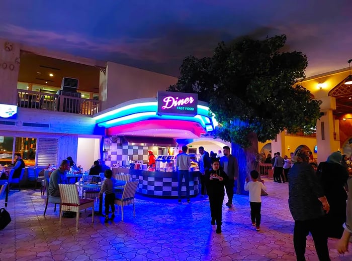 Patrons gather around an indoor kiosk adorned with large neon bars illuminating the ceiling, complemented by a neon sign reading 'Diner Fast Food.'