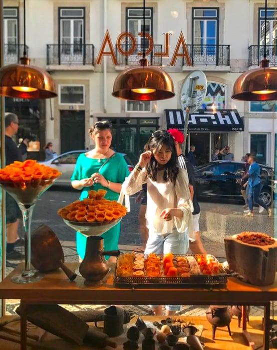 Two women bask in the sunshine outside, admiring the pastry displays illuminated by bronze pendant lamps.