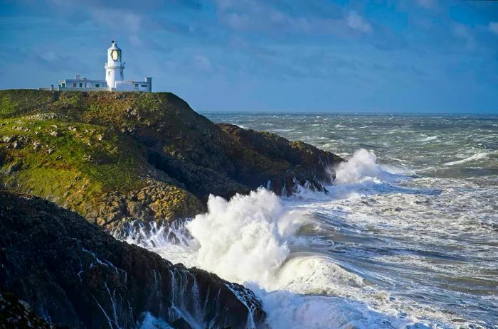 Waves crash against the rugged coastline beneath Strumble Head lighthouse, Wales