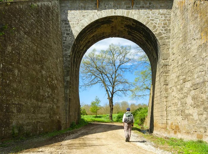 A pilgrim with a backpack strolls through a stone arch along the Via Francigena, near Vetralla.