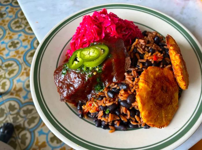 A dish of rice and beans topped with glossy barbecued ribs, crispy tostones, and vibrant pink slaw.