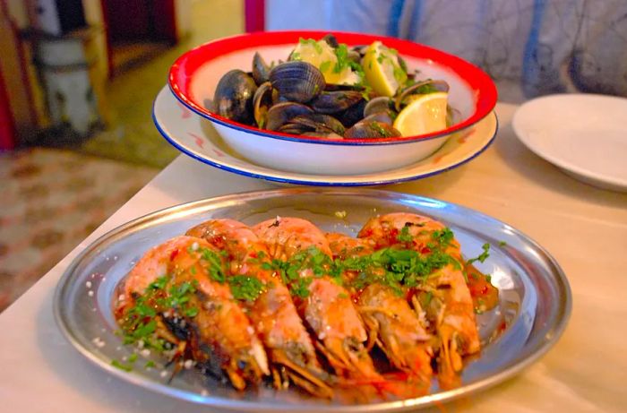 A gleaming silver platter showcases five head-on prawns arranged side by side, each drizzled with a fragrant herb sauce. A bowl of steamed mussels rests in the background.