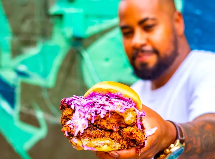 A man showcases a gigantic fried chicken sandwich piled high with purple cabbage slaw.