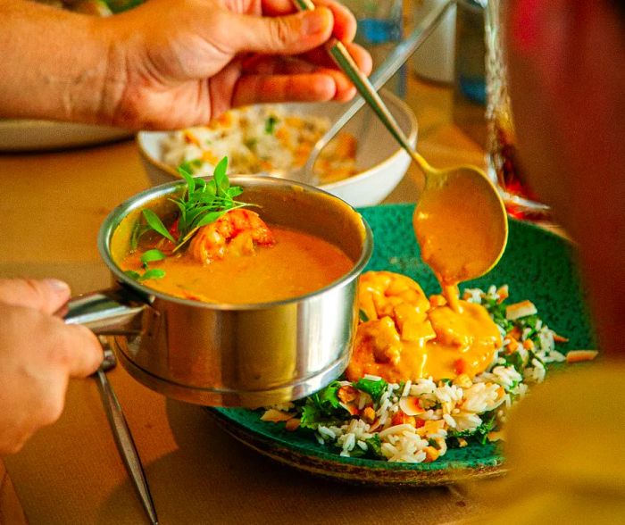 A server pours curry sauce from a pot, with a shrimp floating in it, over a bed of rice and vegetables on a plate.