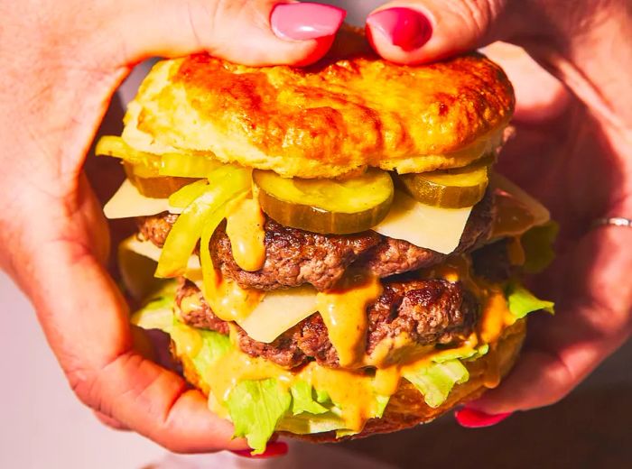 Hands grasp a towering biscuit sandwich filled with multiple burger patties, pickles, cheese, and crisp lettuce.