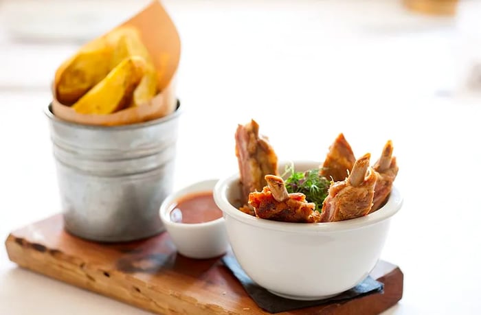 A rustic wood plank displays individual bone-in ribs alongside a tin can filled with french fries and dipping sauce.