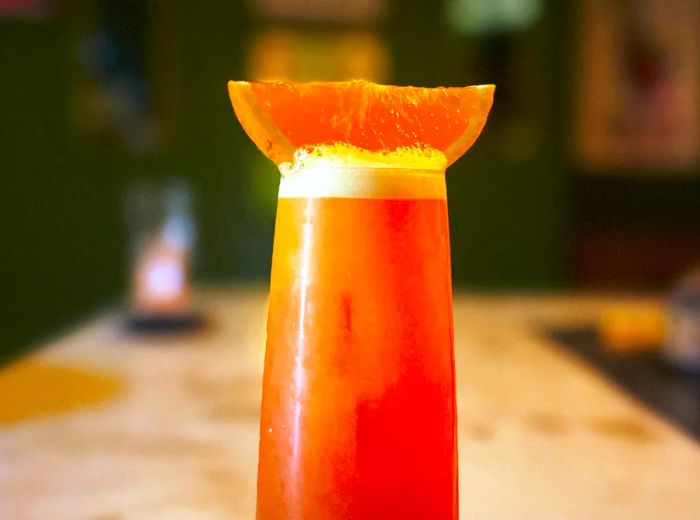 A tall, frothy cocktail garnished with a slice of orange, resting on a bar counter.