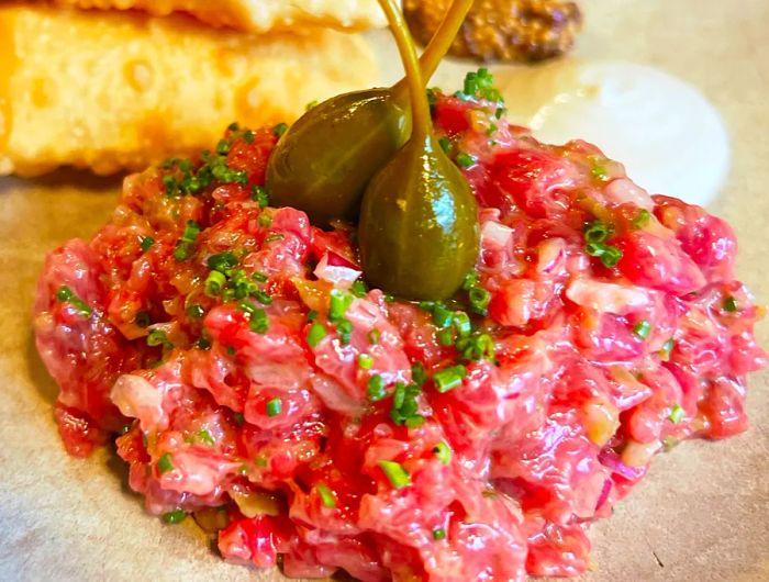 A serving of steak tartare garnished with caper berries, accompanied by bread on the side.