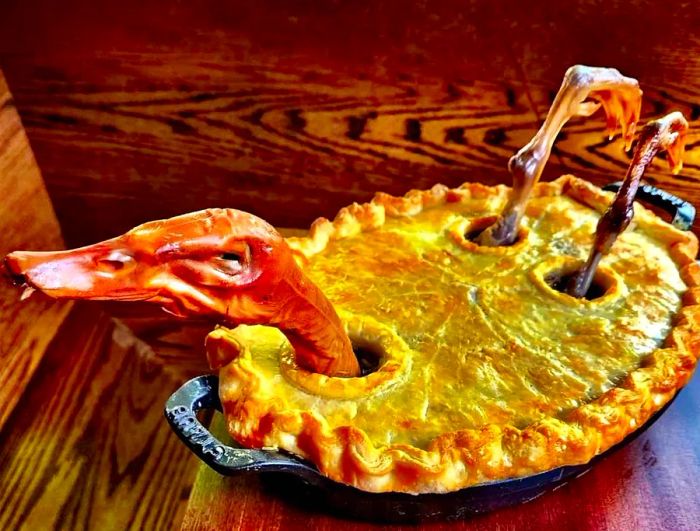 A savory pie featuring the head and legs of a roast duck protruding from the crust.