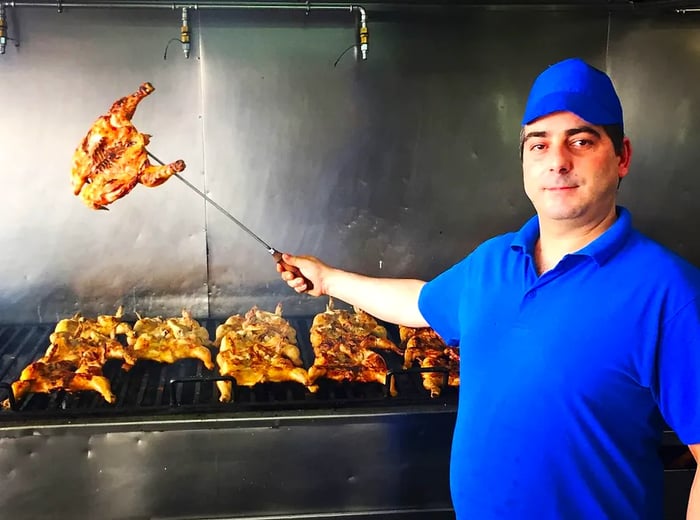 A chef lifts a skewer of roast chicken above a kitchen grill.