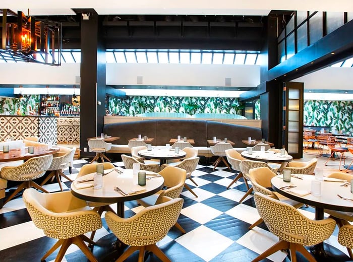 A bright dining area featuring large, round midcentury tables and textured chairs, with checkerboard flooring, glass panels beneath a high ceiling, and a strip of open space leading to lush greenery.