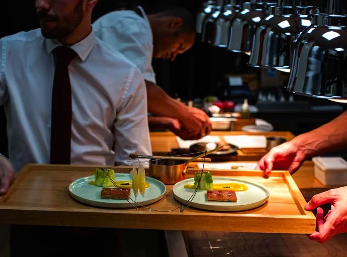 A waiter emerges from a dimly lit kitchen, balancing a tray laden with several dishes and a wire hanger showcasing slices of food.