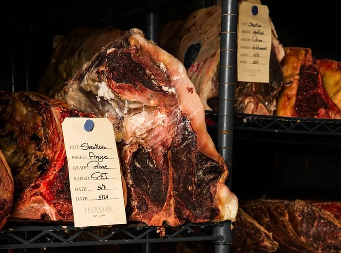 Large cuts of raw meat displayed on metal racks, each with handwritten labels.