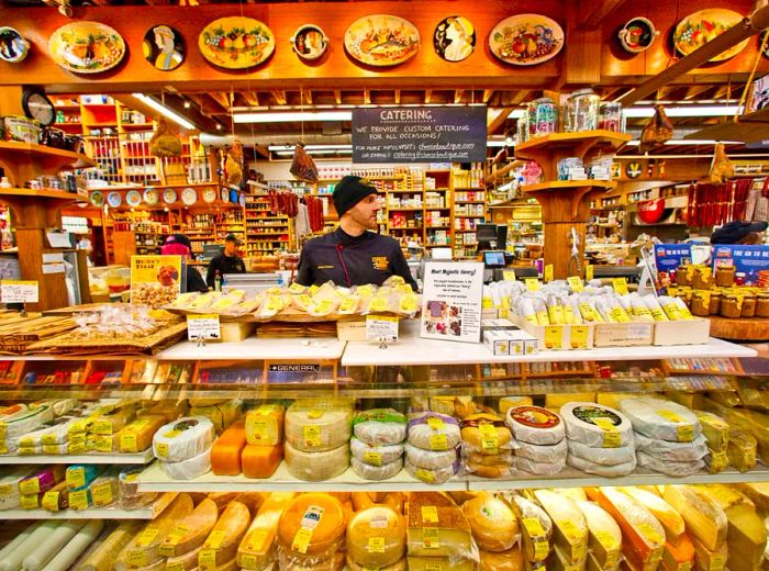 A staff member glances away from the camera as they stand behind a deli case brimming with cheese, surrounded by even more cheese.