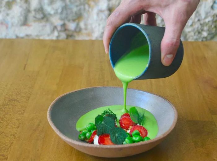 A server elegantly pours vibrant, thick pea soup from a ceramic cup into a bowl already filled with slices of pickled strawberries, walnuts, and cashew cream.