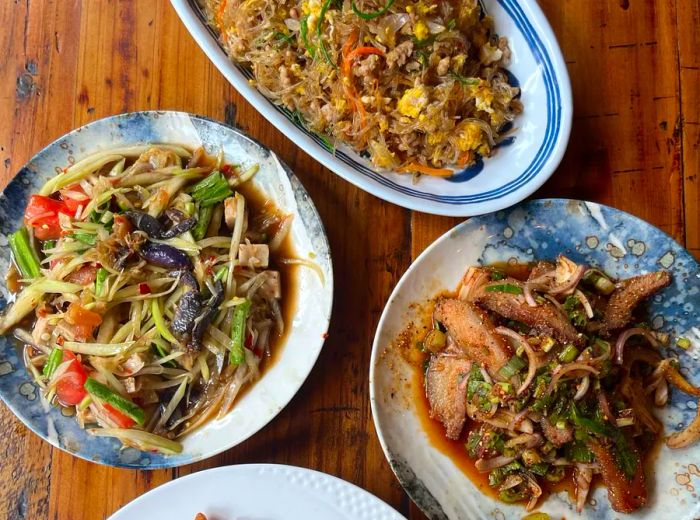 A wooden table displays four dishes, including vibrant papaya salad, fragrant fried rice, and various meat dishes.